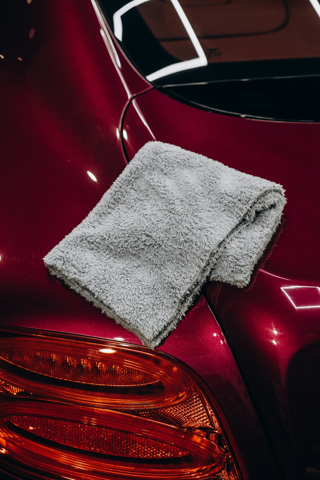 A folded grey plush microfiber towel sits on the gleaming trunk of a modern red car after a professional detail. The flawless, glossy paint reflects the overhead lights, showcasing the results of polishing and paint correction. Image is suitable for themes of automotive care, luxury vehicle maintenance, detailing services, and car cleaning products.