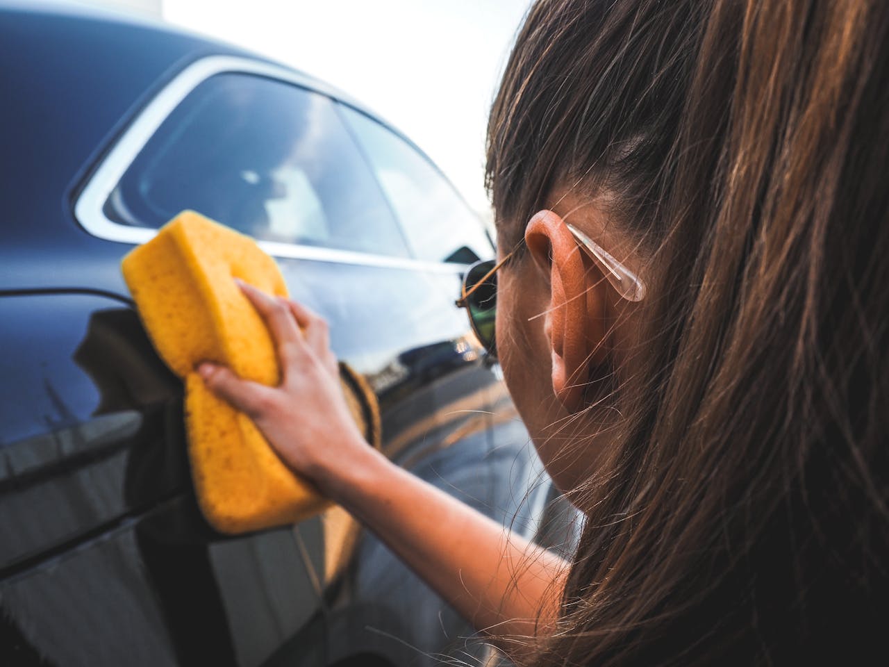 Close-up of a woman handwashing a black car using a yellow sponge, highlighting cleaning process.
